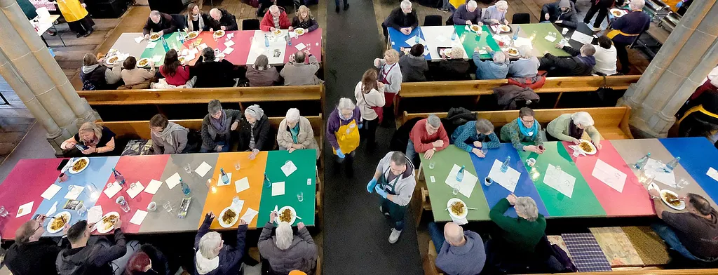 Blick von oben ineinen Kirchraum, in dem Menschen an Bänken sitzen und gemeinsam essen.